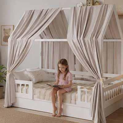 Ciel de lit cabane blanc en bois avec rideaux beiges posée dans une chambre, enfant assise lisant sur le lit cabane
